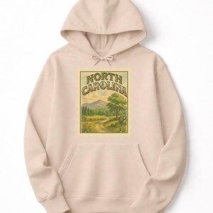 May include: A light beige hoodie with a front pocket and drawstring hood. The front features a rectangular graphic with the words "NORTH CAROLINA" above a landscape scene of a road, trees, and mountains.