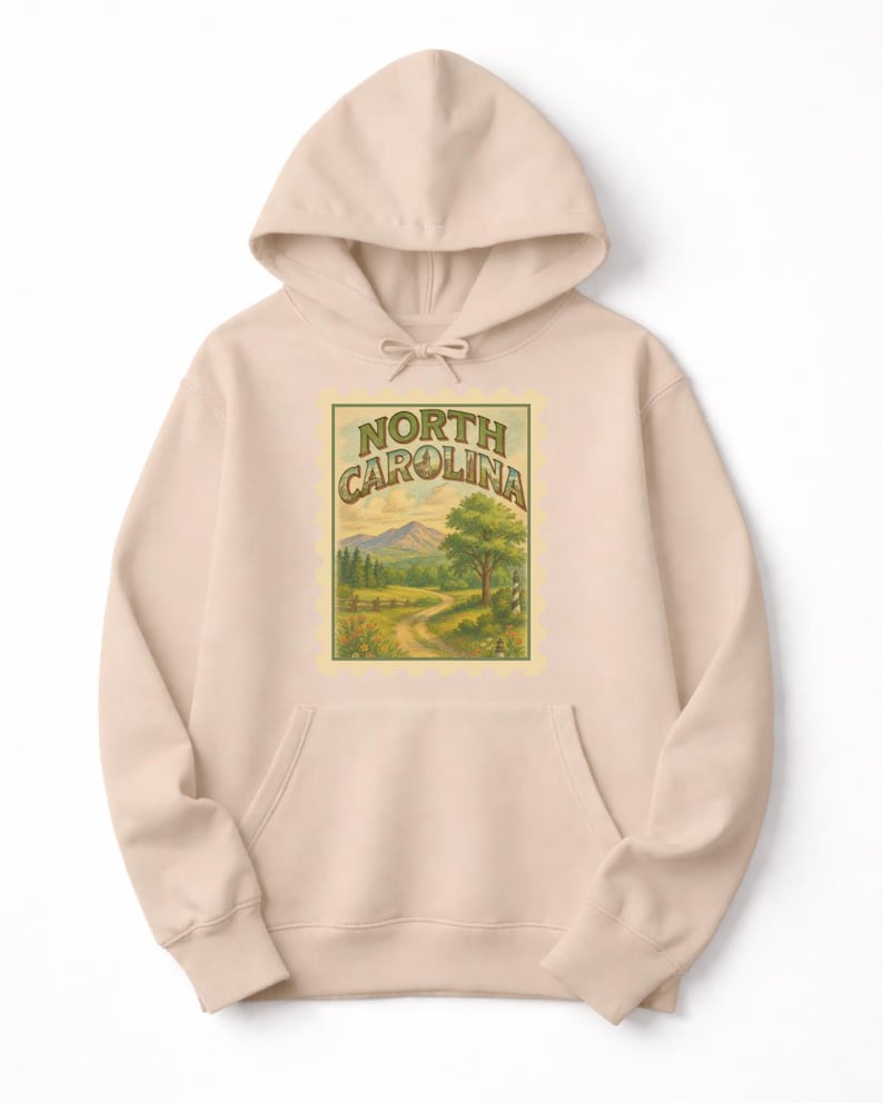 May include: A light beige hoodie with a front pocket and drawstring hood. The front features a rectangular graphic with the words "NORTH CAROLINA" above a landscape scene of a road, trees, and mountains.