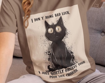 May include: A tan t-shirt featuring a black cat illustration with large eyes and the text "I DON'T BRING BAD LUCK. I JUST QUIETLY OBSERVE YOUR POOR LIFE CHOICES." The cat is sitting with a slightly mischievous expression.