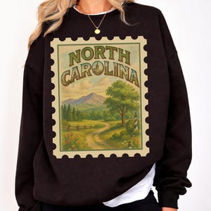 May include: Black sweatshirt featuring a vintage-style North Carolina stamp design. The stamp depicts a scenic landscape with mountains, trees, and a winding road. The words "North Carolina" are arched across the top of the stamp.