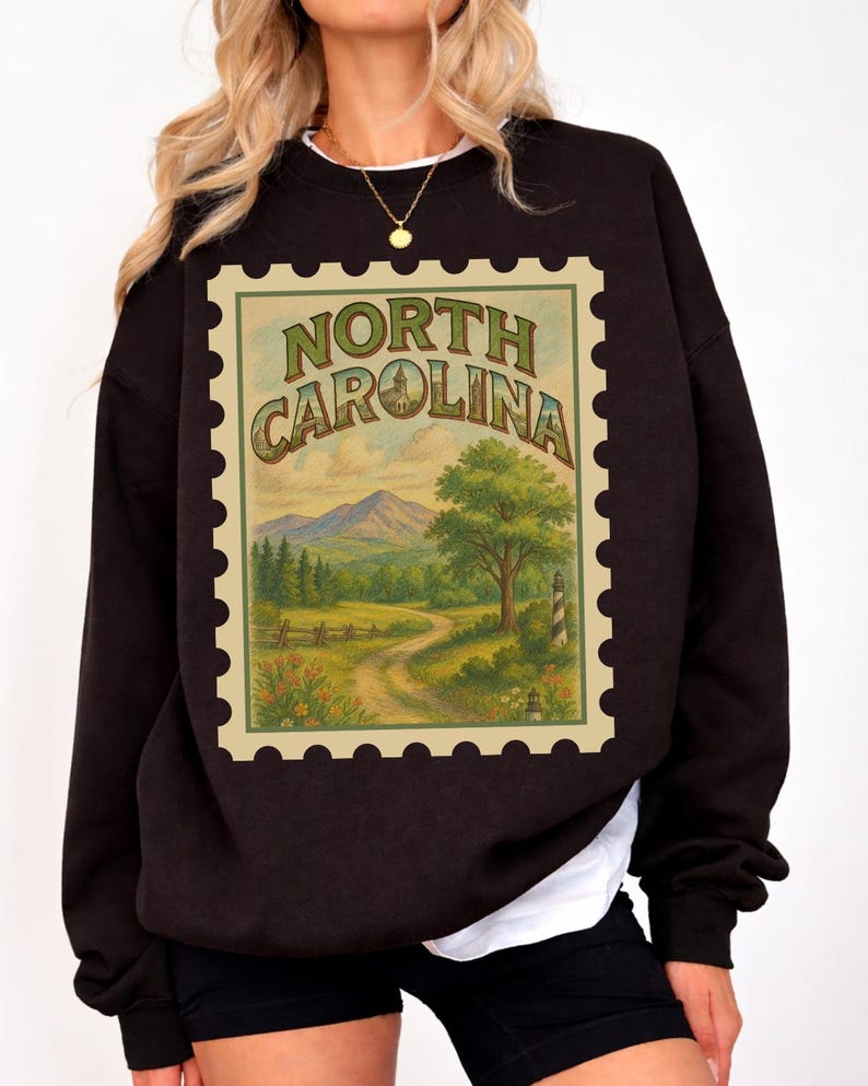May include: Black sweatshirt featuring a vintage-style North Carolina stamp design. The stamp depicts a scenic landscape with mountains, trees, and a winding road. The words "North Carolina" are arched across the top of the stamp.