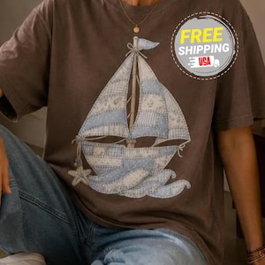 May include: Brown t-shirt with a sailboat design. The sailboat is light blue and white, with a patchwork pattern. The shirt has short sleeves and a round neckline. The image also includes the text "FREE SHIPPING USA".