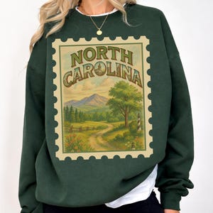May include: Dark green sweatshirt featuring a vintage-style North Carolina stamp design. The stamp includes a scenic landscape with mountains, a winding road, a tree, and a lighthouse. The text "NORTH CAROLINA" is arched across the top of the stamp.