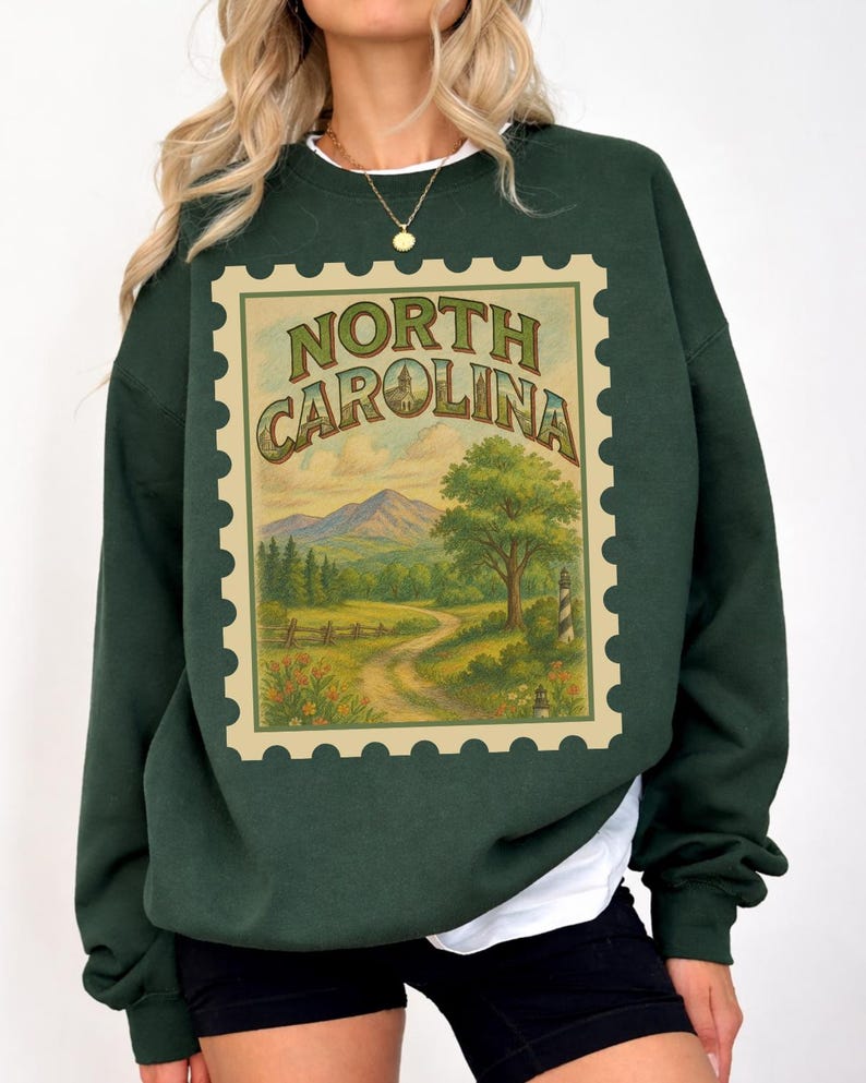 May include: Dark green sweatshirt featuring a vintage-style North Carolina stamp design. The stamp includes a scenic landscape with mountains, a winding road, a tree, and a lighthouse. The text "NORTH CAROLINA" is arched across the top of the stamp.