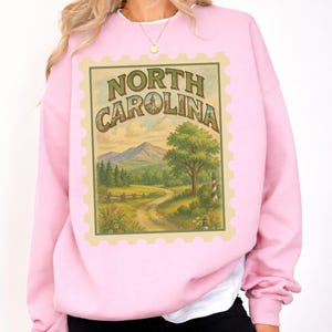 May include: A light pink sweatshirt featuring a North Carolina stamp design. The stamp includes the text "NORTH CAROLINA" and a landscape scene with mountains, trees, and a winding road. The sweatshirt has long sleeves and a crew neck.