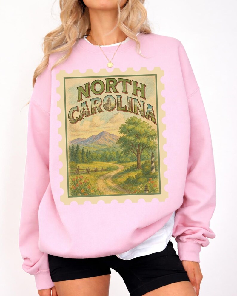 May include: A light pink sweatshirt featuring a North Carolina stamp design. The stamp includes the text "NORTH CAROLINA" and a landscape scene with mountains, trees, and a winding road. The sweatshirt has long sleeves and a crew neck.