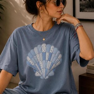 May include: A blue t-shirt featuring a seashell design. The seashell is made of various blue and white patterned fabrics. The shirt has short sleeves and a relaxed fit, suitable for casual wear.