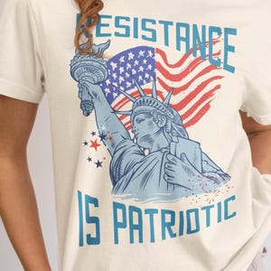 May include: A cream-colored t-shirt featuring a blue graphic of the Statue of Liberty holding a torch in front of a red and white American flag. The text RESISTANCE IS PATRIOTIC is printed in blue block letters above and below the graphic.