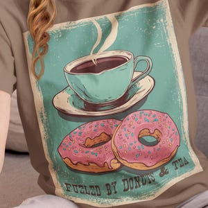 May include: A brown t-shirt featuring a vintage-style graphic of a teal teacup and saucer with steam rising, and two pink donuts with sprinkles. The text "Fueled by Donuts & Tea" is below the image.