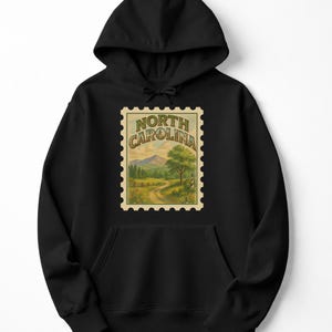 May include: Black hooded sweatshirt featuring a vintage-style North Carolina stamp design. The stamp depicts a scenic landscape with mountains, trees, and a winding road. The text "NORTH CAROLINA" is arched across the top of the stamp.