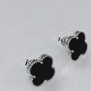 May include: A pair of clover-shaped stud earrings. Each earring features a black center surrounded by a silver-colored beaded border. The earrings are displayed on a light gray surface.