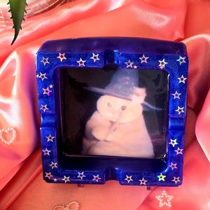 May include: A square, dark blue ashtray with a picture of a cat wearing a wizard hat. The ashtray is decorated with holographic star-shaped accents around the edges. The ashtray is sitting on a pink satin surface.
