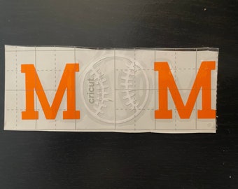 May include: Orange vinyl letters spelling "MOM" with a white baseball graphic in the center. The letters and graphic are on a clear transfer sheet.