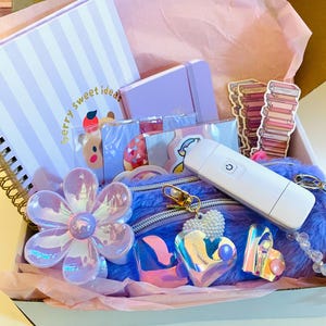 Op de afbeelding: Een geschenkdoos met een spiraalgebonden notitieboek met de tekst berry sweet ideas, een paars dagboek, een pluizig paars tasje, een witte draagbare ventilator, iriserende bloem- en hartvormige accessoires en stickers van boeken.