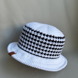 May include: A white crochet bucket hat with a black and white houndstooth pattern around the crown. The hat has a small brim and a brown leather tag attached to the side.