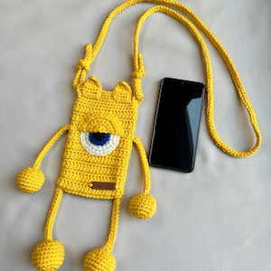 May include: A handmade yellow crochet phone case with a monster design. The case features a large eye with blue and white details, long dangling arms and legs with yellow pom-poms, and a matching strap. A black smartphone rests beside it.