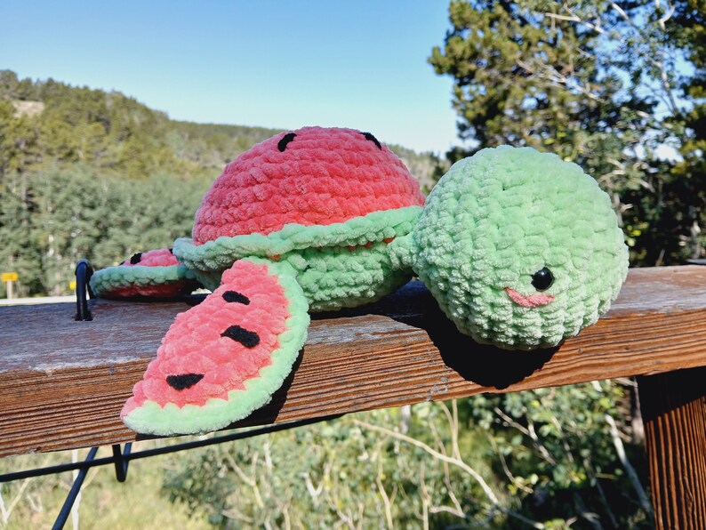 May include: A handmade crochet watermelon plushie with a red top, green rind, and black seed details. The plushie has a smiling face and is placed on a wooden surface. The overall theme is summer and fruit.