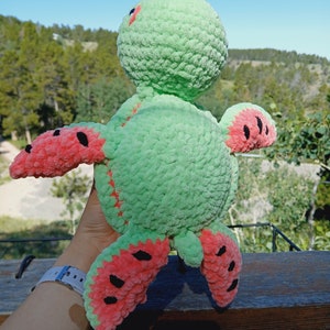 May include: A handmade plush turtle toy in shades of green and red. The turtle's body is a soft green, with red accents on the flippers and head. The flippers are designed to resemble watermelon slices, with black seed details. The toy is made of a textured yarn.