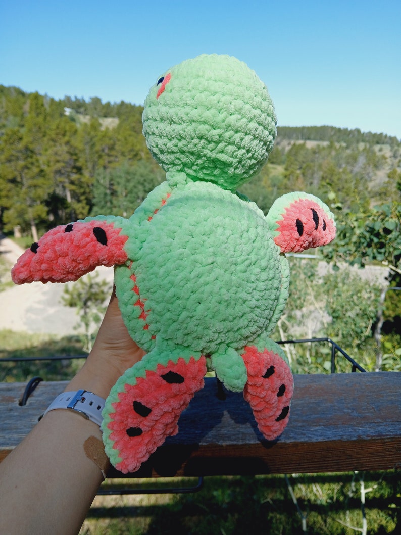May include: A handmade plush turtle toy in shades of green and red. The turtle's body is a soft green, with red accents on the flippers and head. The flippers are designed to resemble watermelon slices, with black seed details. The toy is made of a textured yarn.