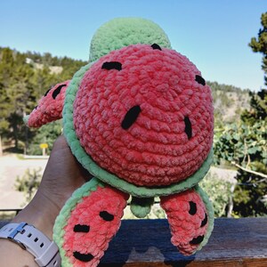 May include: A plush, crocheted turtle toy in the shape of a watermelon. The turtle features a red body with black seed details, green edges, and a green head. The toy is held up against a backdrop of a blue sky and trees.
