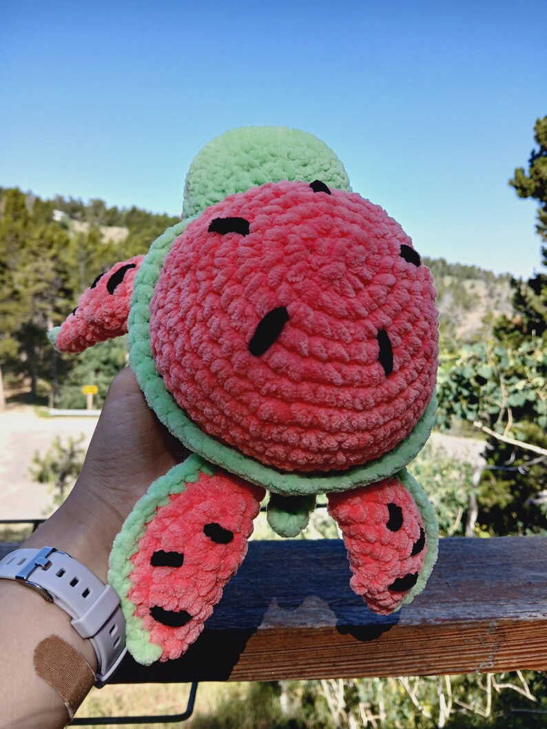 May include: A plush, crocheted turtle toy in the shape of a watermelon. The turtle features a red body with black seed details, green edges, and a green head. The toy is held up against a backdrop of a blue sky and trees.