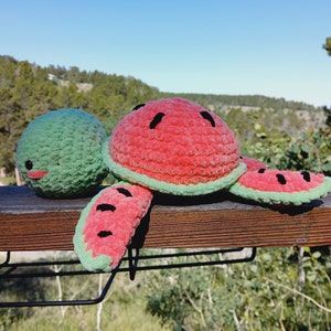 May include: A handmade crochet watermelon turtle plush toy. The turtle has a red shell with black seed details, green edges, and green flippers. The head is green with a smiling face. The plush is on a wooden surface.