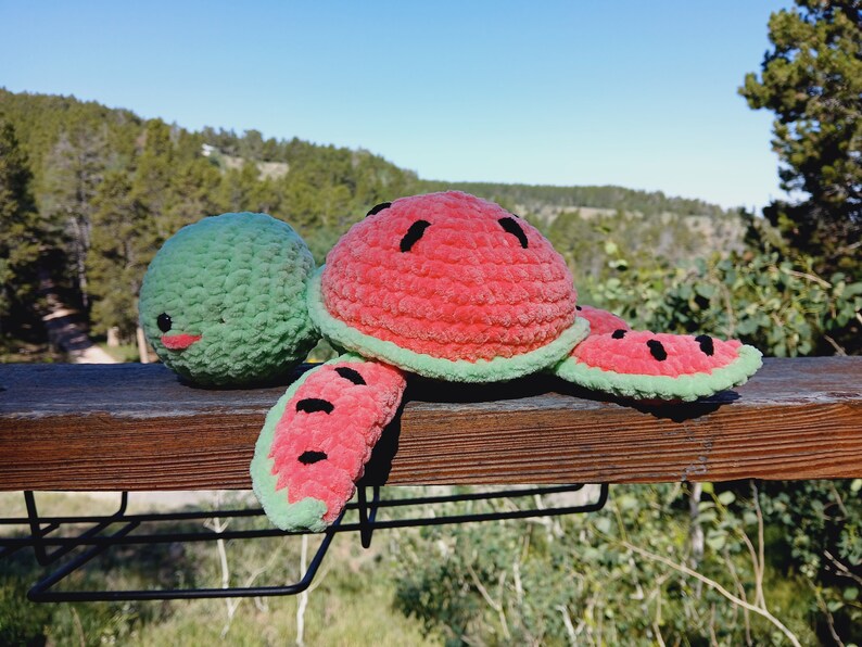 May include: A handmade crochet watermelon turtle plush toy. The turtle has a red shell with black seed details, green edges, and green flippers. The head is green with a smiling face. The plush is on a wooden surface.