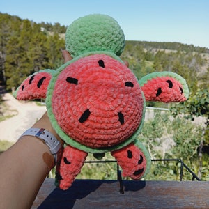 May include: A handmade plush turtle toy with a watermelon design. The turtle's shell is red with black seed details, and the edges and head are green. The toy is held up against a backdrop of trees and a blue sky.
