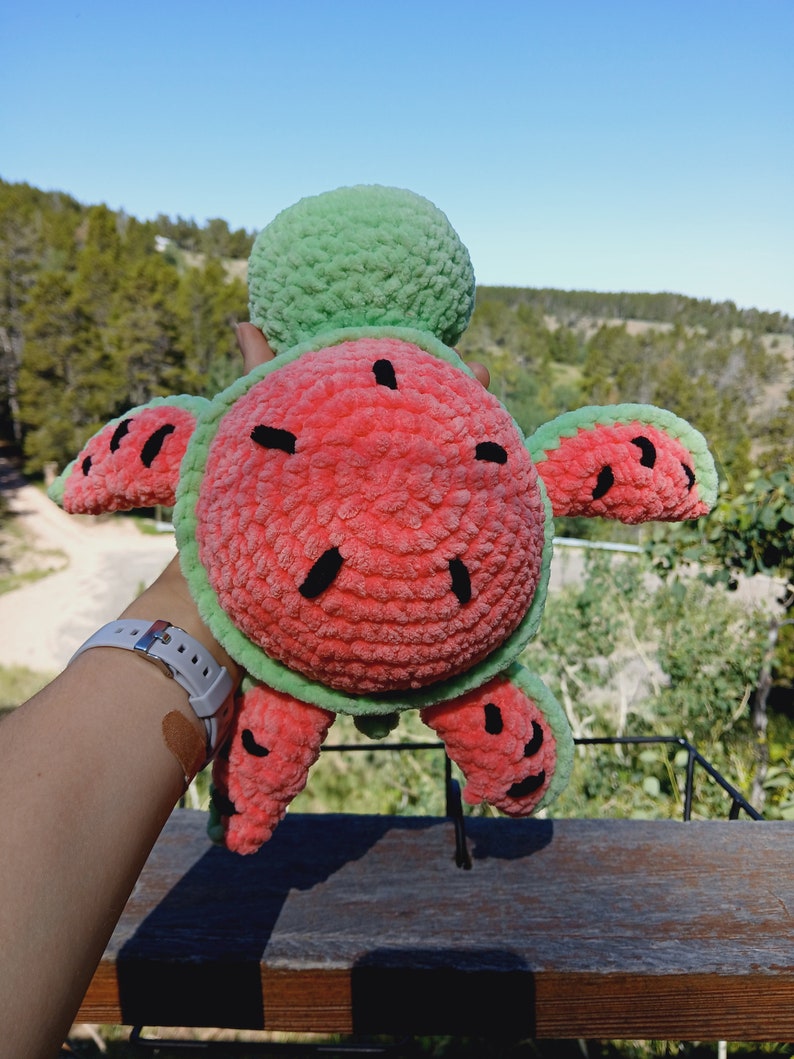 May include: A handmade plush turtle toy with a watermelon design. The turtle's shell is red with black seed details, and the edges and head are green. The toy is held up against a backdrop of trees and a blue sky.
