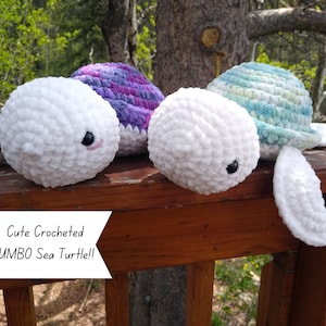 May include: Two crocheted sea turtle plushies, one with a purple and white shell and the other with a blue and white shell. Both have white fluffy bodies and black eyes. The text "Cute Crocheted JUMBO Sea Turtle!!" is on a white sign in front of the turtles.