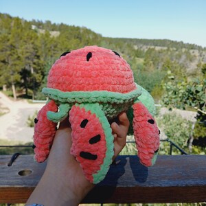 May include: A handmade watermelon-shaped plush toy. The toy features a red body with black seed details and a green rind. The plush is held up against a backdrop of trees and a blue sky.