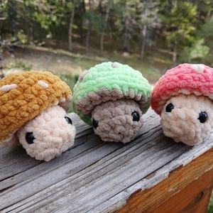May include: Three handmade crochet mushroom figures in shades of yellow, green, and pink. Each mushroom has a beige stem and a textured cap with white spots. The mushrooms are displayed on a weathered wooden surface.