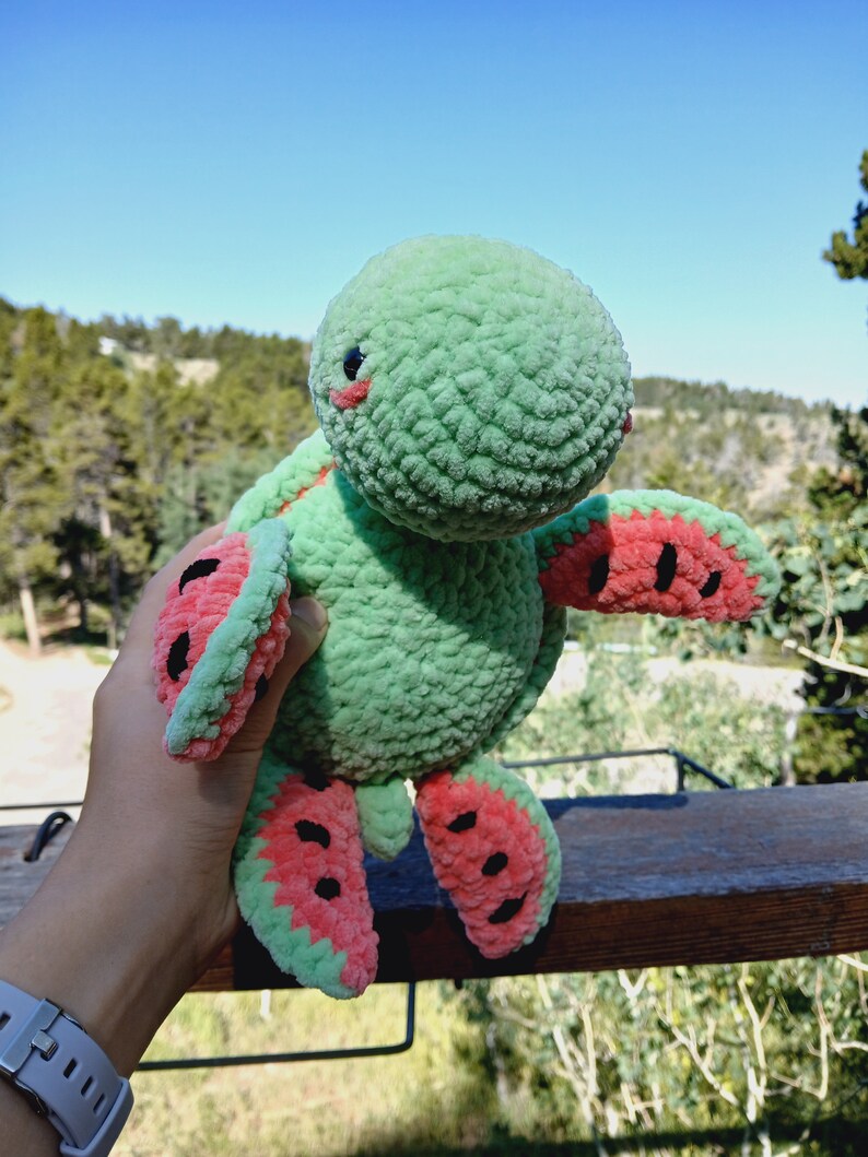 May include: A handmade green plush turtle with watermelon-slice flippers and shell details. The turtle has a round head, small black eyes, and pink cheeks. The plush toy is held up against a backdrop of trees and a blue sky.