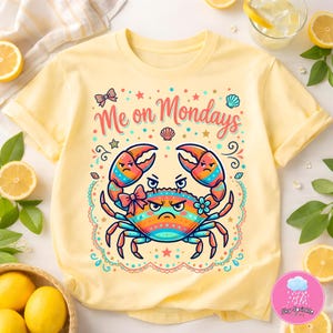 May include: Yellow t-shirt with a crab graphic and the text "Me on Mondays". The crab is colorful with a grumpy expression. The design includes a bow, stars, and a seashell.