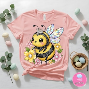 Pode incluir: T-shirt rosa claro com uma abelha de desenho animado usando uma coroa, cercada por flores. A abelha tem listras pretas e amarelas e asas iridescentes. A camiseta é amarrada na cintura e tem decote redondo.