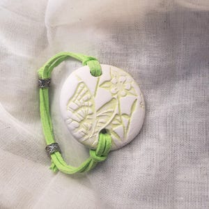 May include: A white ceramic bracelet pendant with a butterfly and floral design, connected to a bright green suede cord. The pendant is round and has a raised relief design. The cord is secured with silver-colored metal beads.