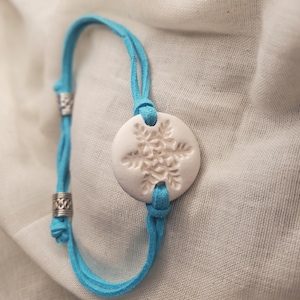 May include: A light blue suede bracelet with a white, circular pendant featuring a snowflake design. The bracelet has silver-colored metal accents and is tied with knots. The pendant is approximately 1 inch in diameter.