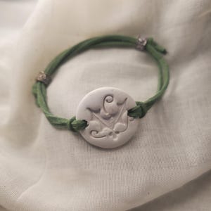 May include: A handmade bracelet featuring a white, circular pendant with an embossed floral design. The pendant is attached to a green suede cord. The bracelet has silver-toned metal accents and is displayed on a white fabric background.