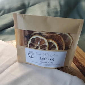 May include: A brown paper bag with a clear window, containing dried lemon and orange slices, cinnamon sticks, and other botanicals. The bag has a label that reads "Let's Go! Simmer Pot Kit" with the brand name "Mindful RN Creations".