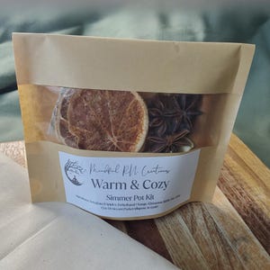 May include: A brown paper bag containing a simmer pot kit. The kit includes dried orange slices, star anise, and other spices. The label reads "Mindful RN Creations Warm & Cozy Simmer Pot Kit."