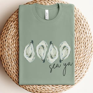 May include: Sage green t-shirt with four oyster illustrations and the handwritten text "sea ya". The shirt is made of a soft material and is displayed on a woven, circular surface.