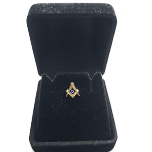 Pode incluir: Um alfinete de lapela em tom dourado com o símbolo maçónico do esquadro e compasso, com um centro em esmalte azul e a letra G, apresentado dentro de uma caixa de joias forrada a veludo preto com uma tampa articulada aberta.