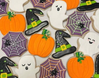May include: A selection of Halloween-themed biscuits. The biscuits feature orange pumpkins, white ghosts, black witch hats with green details, and purple spiderwebs with black spiders. The biscuits are decorated with icing and are arranged on a white surface.