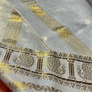 May include: Close-up of a white textile with gold decorative borders. The borders feature intricate patterns, including peacock and tree motifs. The fabric is folded, showcasing the detailed design and texture.