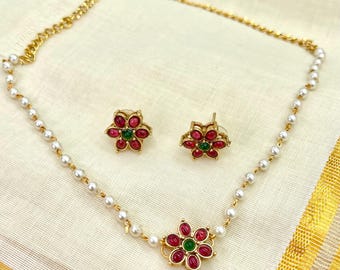 May include: A gold-toned necklace and earring set. The necklace features a strand of small white pearls and a gold chain. The pendant and earrings are flower-shaped, with red and green gemstone accents. The jewelry set is displayed on a cream-colored fabric.