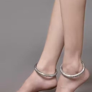 May include: A pair of silver anklets with an engraved pattern. The anklets are worn around the ankles, and the image shows the feet and lower legs against a gray background. The anklets appear to be made of metal.