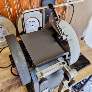 Puede incluir: Una máquina de afilar gris y negra con dos muelas. La máquina tiene un brazo de metal con un mango negro y una placa de metal en la parte superior. Las muelas están etiquetadas con "DC-250".