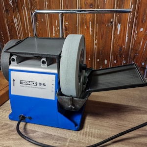 May include: A Tormek T-4 water-cooled sharpening system. The machine has a blue base with a gray grinding wheel and a black tray. The text "Tormek T-4 Water Cooled Sharpening System" is printed on the front.