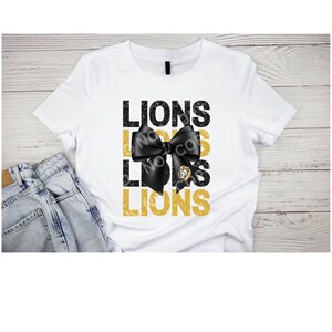 May include: White t-shirt with a black bow and gold glitter text that reads "Lions" three times.