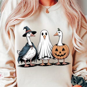 Op de afbeelding: Een beige sweatshirt met een Halloween-ontwerp. De afbeelding toont drie ganzen verkleed als heks, spook en pompoen. De cartoon-stijl illustratie is perfect voor het herfstseizoen.